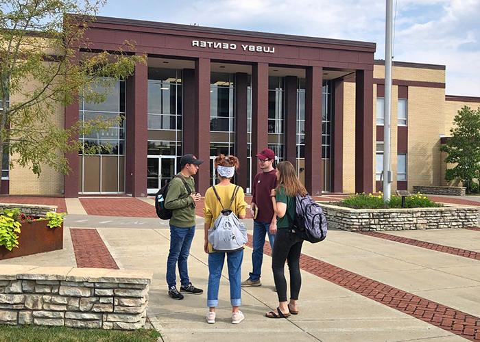 Four students with backpacks stand and talk in front of Lusby Center, a modern brick building with large windows.